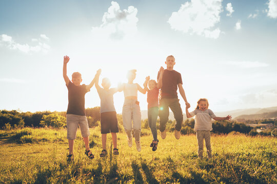 Six kids brothers and sisters teenagers and little kids funny jumping holding hands in hands on the green grass meadow with an evening sunset background light. Happiness and careless childhood concept