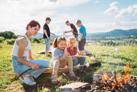 Three sisters sitting near campfire, smiling and roasting marshmallows and candies on sticks over while brothers set up the green tent. Happy family outdoor picnic camping activities concept.