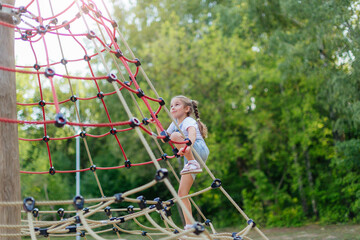 A happy little girl is climbing on an outdoor playground on a summer day. Blonde girl climbs to the top of the game complex