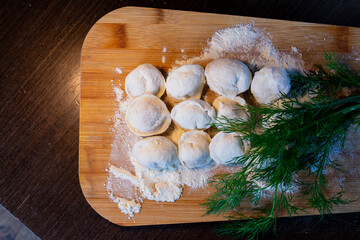 Raw Russian dumplings on a cutting board and ingredients for homemade dumplings on a wooden table, bunches of green dill. The process of making dumplings, ravioli or dumplings with meat.