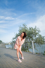 Woman in pink dress standing on river bank with hair flying on wind, blue sky background.