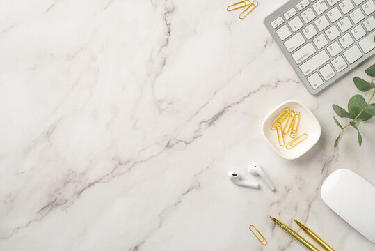 Business concept. Top view photo of workstation keyboard computer mouse wireless earbuds gold pens clips and eucalyptus on white marble background with empty space