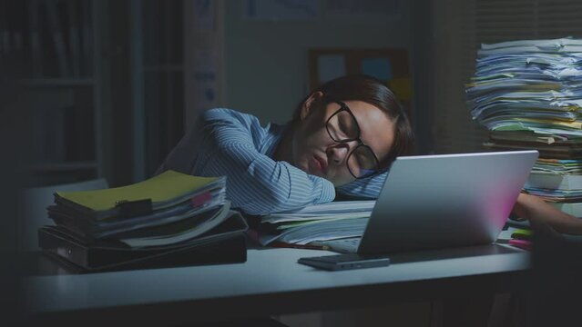 Overworked young Asian office employee wearing eyeglasses sleeping while working on laptop computer overtime in office at night