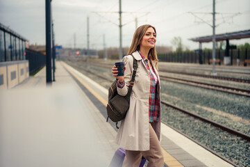 Obraz premium Woman with suitcase waiting for her train on platform of railway station. She is smiling and holding her cup of coffee.