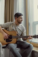 Obraz premium Modern romantic guy musician playing guitar music. Guitarist dreaming, performing musical melody, sitting near window