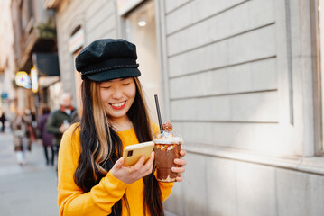 Happy young Asian woman walking down a street in a European city while having a chocolate milkshake and holding her phone