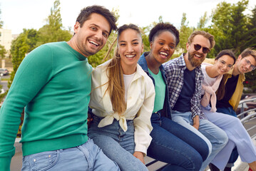Fototapeta premium Diverse group of happy young people having fun during a meet up in the city. Bunch of joyful excited international friends sitting all together on a terrace railing, looking at the camera and smiling