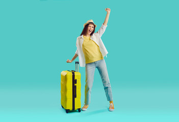 Very happy cheerful excited young woman in casual jeans, T shirt and panama hat standing with yellow suitcase on turquoise background and yelling 'Hooray finally I'm going to travel on summer holiday'