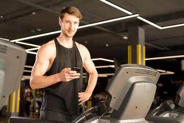 Fototapeta na wymiar Young male athlete standing and using his smartphone before training on treadmill in gym