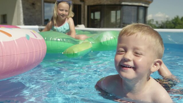 CLOSE UP: Smiling little boy floating in backyard swimming pool learning to swim. Cheerful toddler at his first swimming swings in home pool. Refreshing summer family activities on hot summer days.