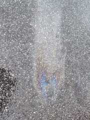 Wet asphalt with stains from gasoline. Dark gray asphalt interspersed with small stones, wet after rain. A rainbow chemical stain from spilled gasoline spread over it.
