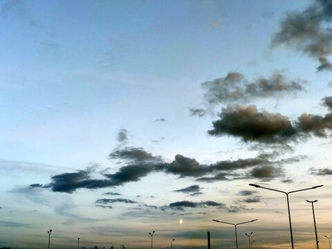 Light Blue Sky With Clouds. Above High Lanterns Under A Light Blue Sky, Rare Cumulus Clouds Of White Gray Hang Low. A Crescent Moon Hangs Low On The Horizon.