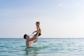 Dad and son are playing in the sea. The father tosses the son up.
