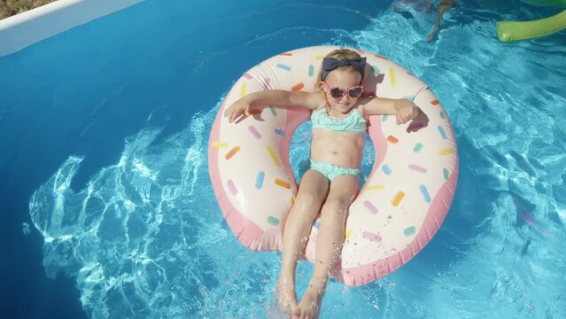 TOP DOWN: Adorable girl with sunglasses on a floatie donut in blue swimming pool. Cute girl having fun floating and making water splashes with her legs. Cheerful kid enjoying summer in backyard pool.