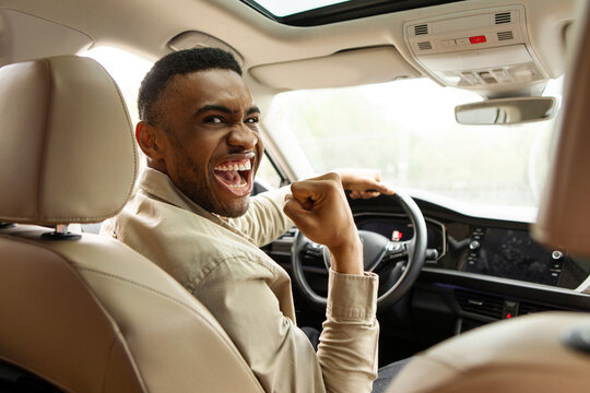 Joyful african american man holding steering wheel shaking fist while sitting in car at car dealership