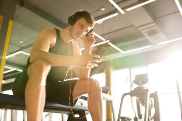 Young sportsman sitting on bench in health club and listening to music online using his smartphone