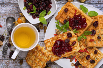 Fresh morning breakfast of baked homemade waffles with berry jam and mint leaves on a white dish on a light wooden table with a cup of green tea with a teaspoon, with a berry in a white cup.
