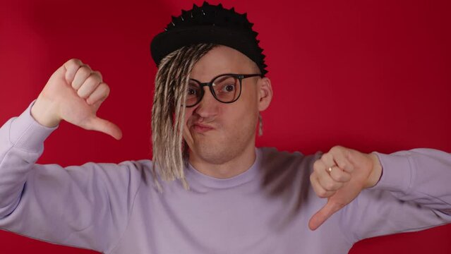 Young handsome man in black cap, glasses showing thumbs down, looking at camera. Stylish guy with blonde dreadlocks showing gesture of dislike on red background.