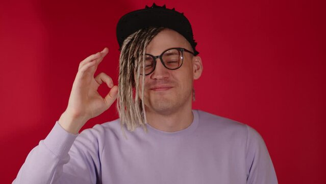 Young handsome man in black cap, glasses showing gesture of OK. Stylish guy with blonde dreadlocks smiling, looking at camera, showing sign of OKAY on red background.