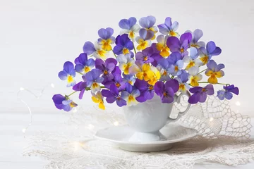 Poster A bouquet of blue, violet, yellow pansies viola in a cup and saucer, a lace napkin on a white table against the wall. Romantic postcard, blur, selective focus. © tachinskamarina