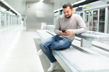 Positive relaxed young bearded man sitting with cellphone on bench on subway platform while waiting for train