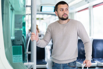 Fototapeta na wymiar Young bearded man traveler riding in modern streetcar, standing leaning on suitcase and holding on to handrails ..