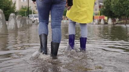 girls in raincoats and rubber boots walk along road flooded with torrential rains, their feet walk through puddles city, splashing water to the sides, the flood is on street, car is driving on water