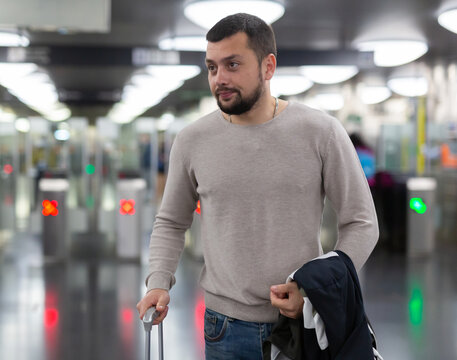 Positive young man going on trip, walking with suitcase to train in subway on blurred background of modern electronic turnstiles..