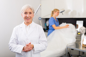 Obraz premium Portrait of smiling confident professional elderly woman cosmetologist wearing white uniform standing in modern medical aesthetic office..