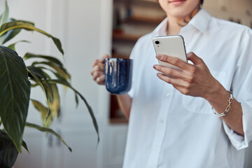 Happy Asian guy typing on phone while holding cup of hot drink