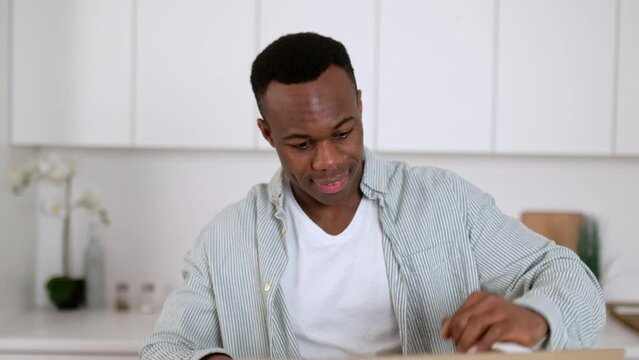 Happy Attractive African American Guy Unpacks His Long Awaited Package, Examines The Clothes That He Bought Online, Stands In Kitchen, Satisfied With The Purchase. Online Shopping, Delivery Concept