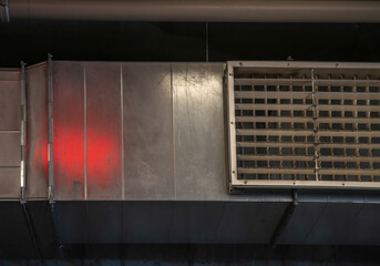 A rectangular sheet metal heating and cooling duct hanging from a ceiling with a metal grille, red light from exit sign reflected on metal