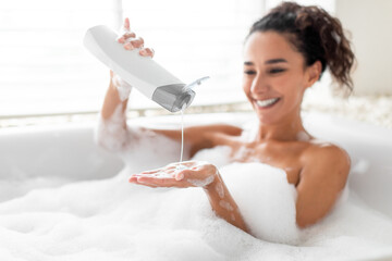 Beautiful young woman taking bubble bath, pouring shampoo into her hand, enjoying home spa procedure, selective focus - obrazy, fototapety, plakaty