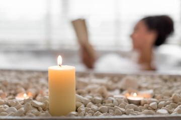 Young woman lying in foamy bath, reading book in relaxing atmosphere, selective focus on candles and stones