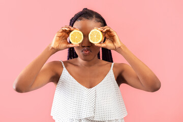 Young black woman with braids holding lemon halves near her eyes, puckering lips on pink studio background