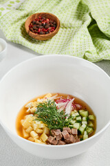 Traditional russian soup - okroshka with fresh vegetables, meat and kvass. Cold summer soup on white table. Aesthetic composition with traditional russian food and spices, textile, spoon.