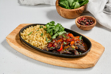 Eastern cuisine - roasted lamb fillet with vegetables and bulgur. Sliced lamb fillet roasted on skillet. Meat dish from lamb fillet with bulgur in skillet on wooden board.