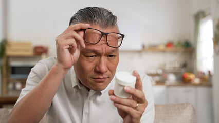 closeup of asian elderly male with presbyopia holding and looking at a pill container with confusion. He squints his eyes trying to see the instruction on bottle clearly - obrazy, fototapety, plakaty