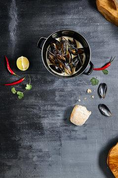 Mussel stew pot with creamy sauce, baguette on stone table. Mussels soup pot contemporary style. Aesthetic composition with seafood. Gastronomy mussel pot dinner concept. Stone table and wooden tray.