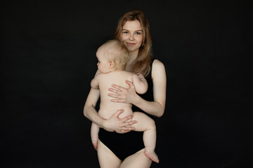 Portrait of young mother in bodysuit holding and embracing naked baby from back on black background. Happy parent enjoying motherhood. Mom and little child relations. Family and baby care concept