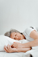 Obraz na płótnie Canvas Mature woman sleeping in comfortable bed, relaxing in bedroom. Elderly caucasian woman enjoying morning dream, rest. Vertical view, copy space