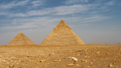 Fototapeta na wymiar The great pyramids of Cheops and Chephren against the blue sky and clouds. The stones are scattered on the sandy soil of the Giza plateau. Silhouettes of Cairo buildings in the distance. Egypt