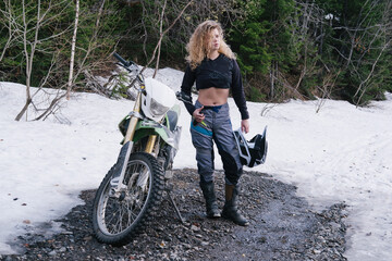 Beautiful young woman standing with helmet near her dirt motorcycle. Snow in summer day, moto...