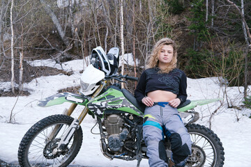 Beautiful young woman standing near her dirt motorcycle. Snow in summer day, moto offroad in...