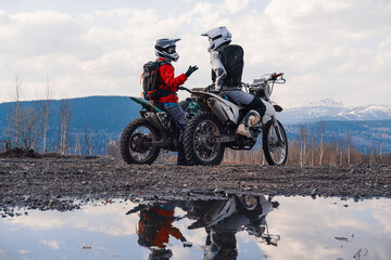 Females wearing helmets and motorcycle gear sitting on motocross motorcycles discuss the journey. Reflection in puddles, snowy peaks on horizon. Offroad travel