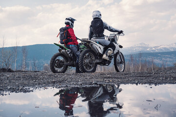 Females wearing helmets and motorcycle gear sitting on dirt motorcycles. Reflection in puddles, snowy peaks on horizon. Offroad travel - obrazy, fototapety, plakaty