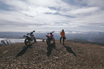Female motorcyclist holding helmet standing with her enduro motorcycle on mountain top, snow peaks...