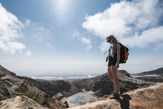 Female traveler wearing sun glasses and backpack walking on crater of volcano above blue lake. Gorely volcano, Kamchatka