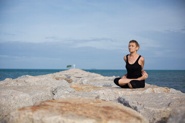 Naklejka na ściany i meble Elderly female sitting in closed lotus position meditating with closed eyes on blue sea beach