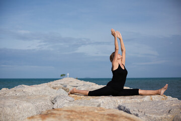 Elderly woman 70 years old doing yoga pose twine on rocky seashore